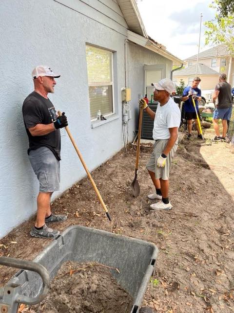 People with shovels and a wheelbarrow working on a yard beside a house