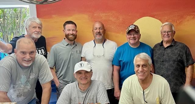 A group of men smiling together in front of a painted sunset mural